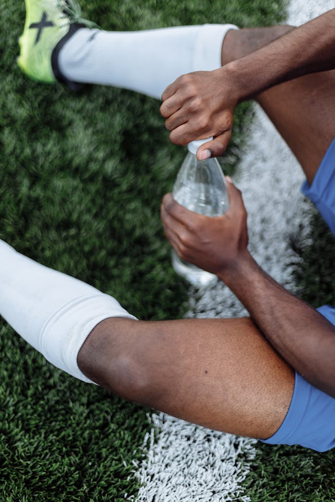 A football player sitting on grass, holding a water bottle while resting on the field.